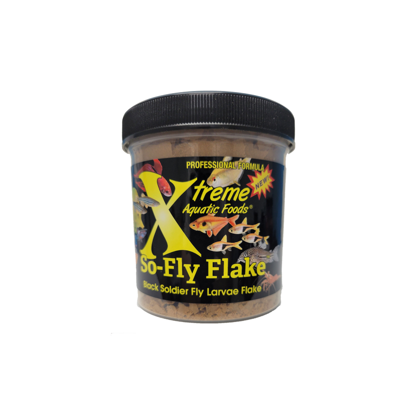 A clear plastic jar containing Xtreme 28g So-Fly Flake fish food, with a black lid and a label featuring the product name and branding with various different tropical fish.